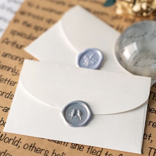Close-up of two white envelopes sealed with silver wax stickers featuring embossed floral designs, set against a background of handwritten text and a glass marble.