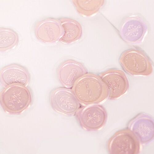 Soft pink and lavender sealing wax stickers with embossed floral and word patterns are scattered on a white surface, highlighting their delicate design.