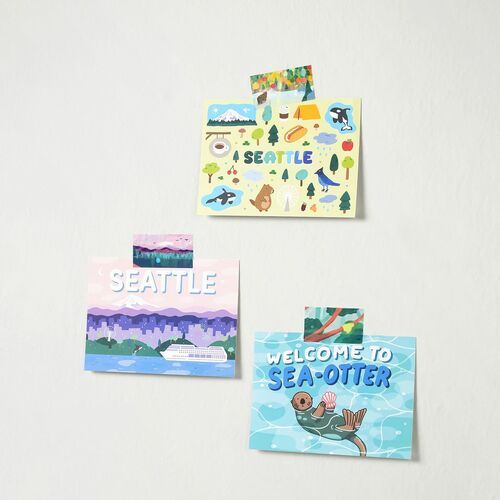 Colorful, playful stickers with vibrant illustrations of Seattle landmarks, nature, and whimsical characters, arranged on a plain white wall.