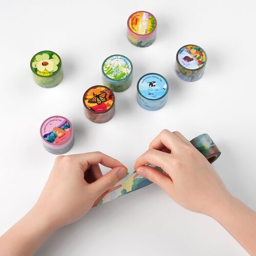 Hands peel back a Seattle scenery masking tape, revealing its colorful, nature-inspired design amidst a display of other themed tapes.