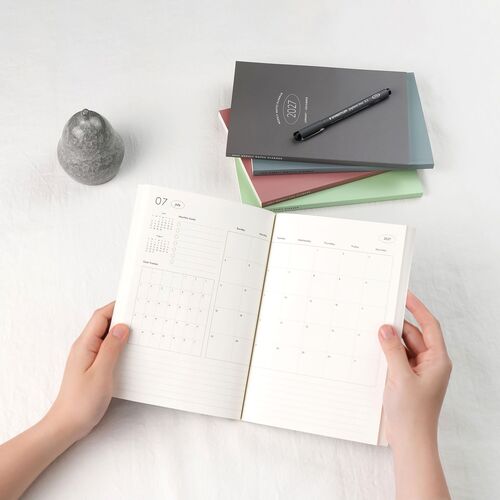 Open planner displaying a clean, minimal layout with soft, natural lighting, complemented by stacked colorful planners and a textured gray candle.