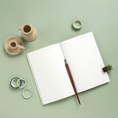 Open grid notebook with a wooden pen on a pastel green surface, surrounded by minimalist ceramic vases and decorative tape rolls.