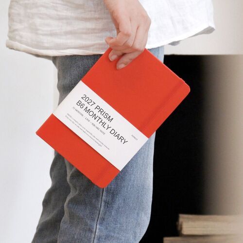 A Tomato Red planner is held in one hand, showing its compact B6 format and secure band around the cover.