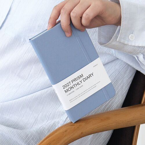 Milky Blue is photographed in a relaxed chair setting, highlighting the rounded hardcover corners and matching elastic band.