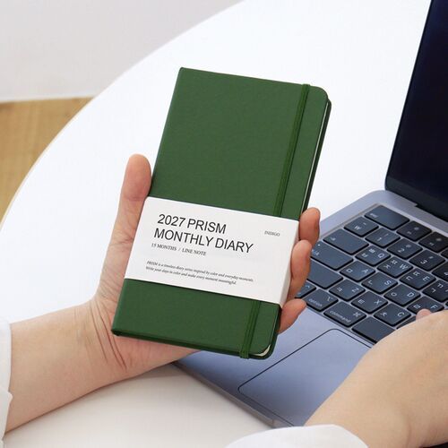 A Tomato Red planner stands in front of several color options, with an open monthly layout and pen in the foreground.
