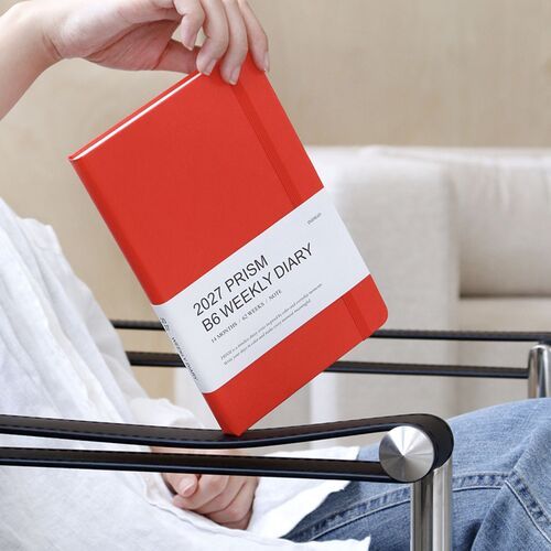 A Tomato Red planner is held at an angle, highlighting its textured hardcover, rounded corners, and coordinating elastic closure.