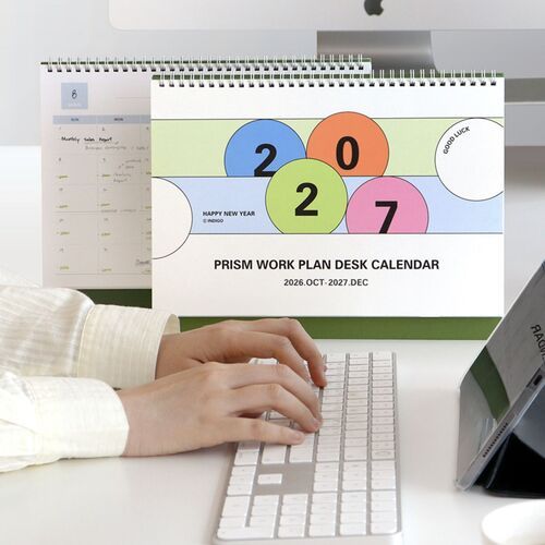 The Prism calendar sits beneath a desktop monitor, showing its colorful geometric cover and green stand in a working setup.