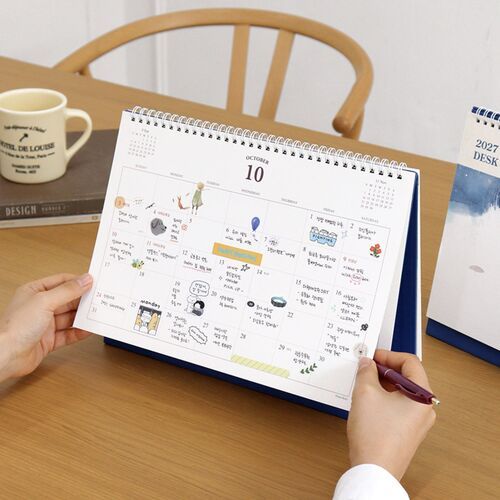 Hands hold an October calendar page above the blue stand, revealing the spacious monthly grid and decorative illustrations.