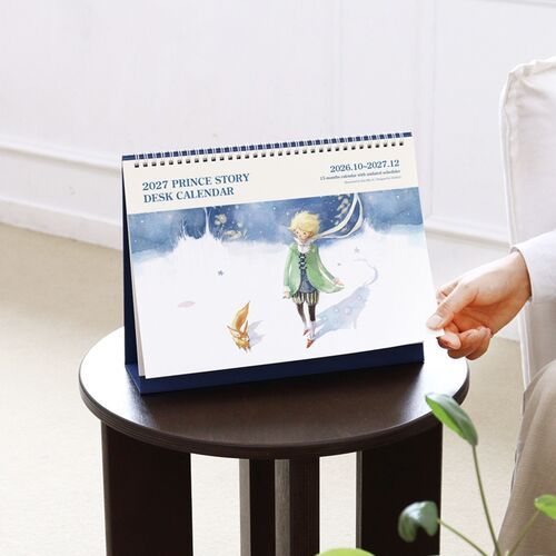 A hand lifts the front page slightly, revealing the thick paper, spiral binding, and blue folded support.