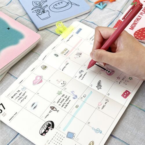 An open monthly spread shows large dated boxes, colorful illustrations, handwritten notes, and a pen in use.