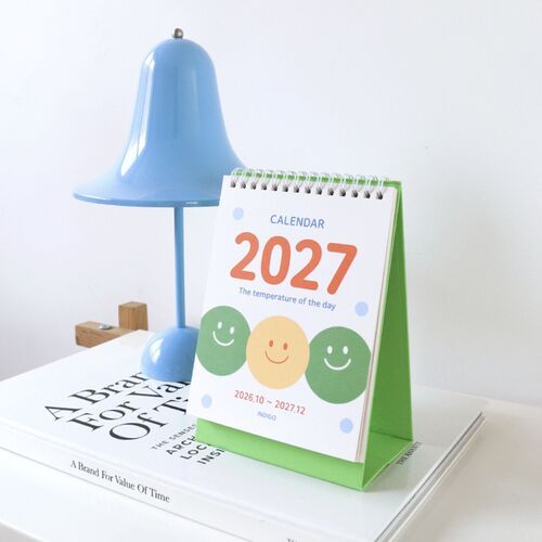 Photo 4: The Pop Orange calendar stands against striped upholstery, highlighting its pale green cover, orange base, and large 2027 lettering.