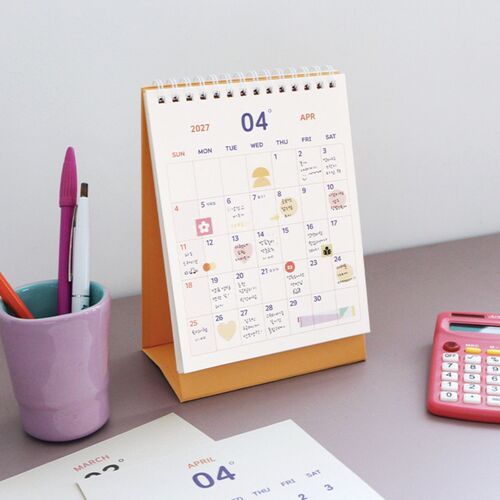 Photo 19: A hand writes on an open calendar page while two standing calendars in Smile Green and Pop Orange sit behind it.