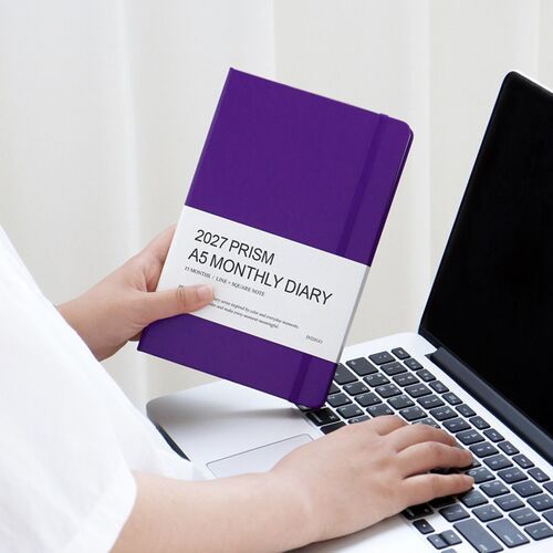 A Sunset Purple planner is held beside a laptop, showing its hardcover format and elastic band while planning at a desk.