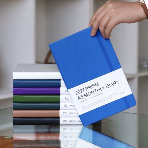The Classic Blue planner is lifted in front of stacked covers, showing its elastic band and rounded hardcover corners.