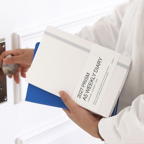 An open planner sits on a wooden desk while a hand writes across its spacious dated weekly layout.