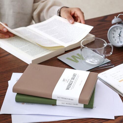 A blue scheduler is held upright against a bookshelf, showing its clean cover and embossed year.