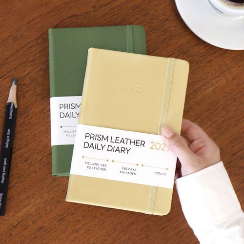 An open daily spread shows time-lined writing areas beside a green pen and a warm desk setting.