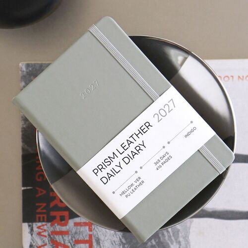 Ink Mellow is held near a white wall, showing its dark cover, rounded corners, and understated 2027 marking.