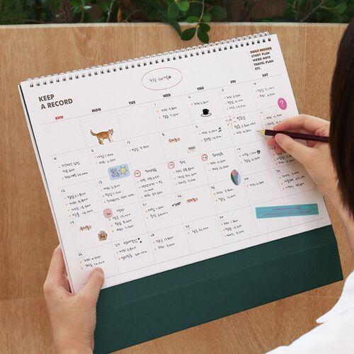 The calendar rests against a dark chair with a coffee cup nearby, giving a sense of its practical tabletop presence.