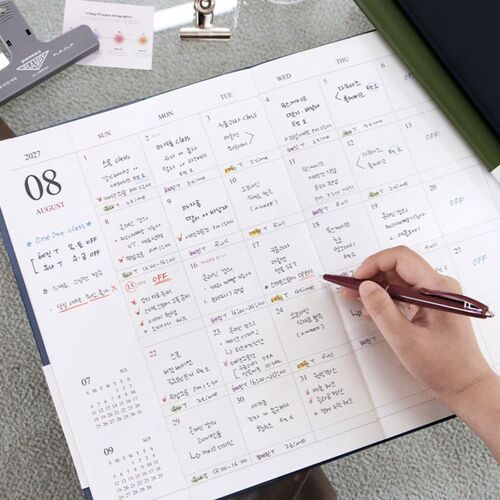 An open scheduler displays a clear PVC flap pocket holding loose papers and reference materials inside the cover.