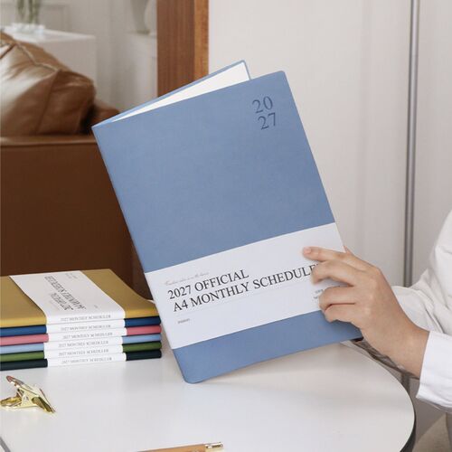 A Navy Blue scheduler stands on a desk beside stationery, presenting the cover and embossed 2027 detail.