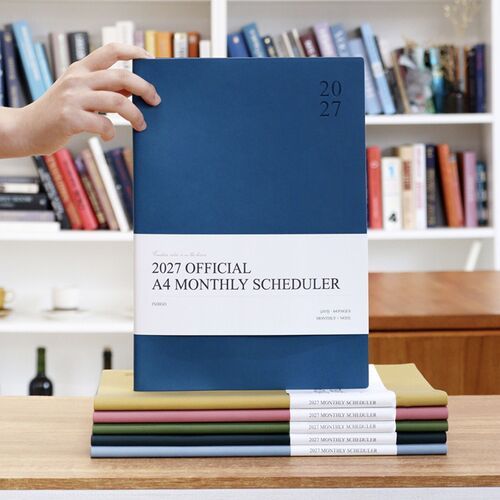 A group of schedulers is stacked on a desk, with Navy Blue, Mustard, Dusty Rose, Olive, and Powder Blue covers visible.