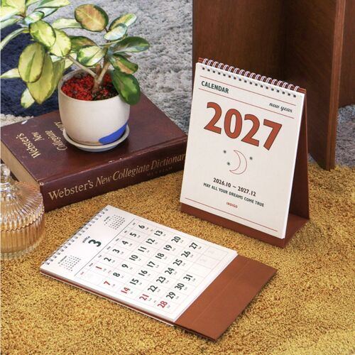 Placed beside books and a potted plant, the calendar adds a practical planning accent to a warm reading corner.