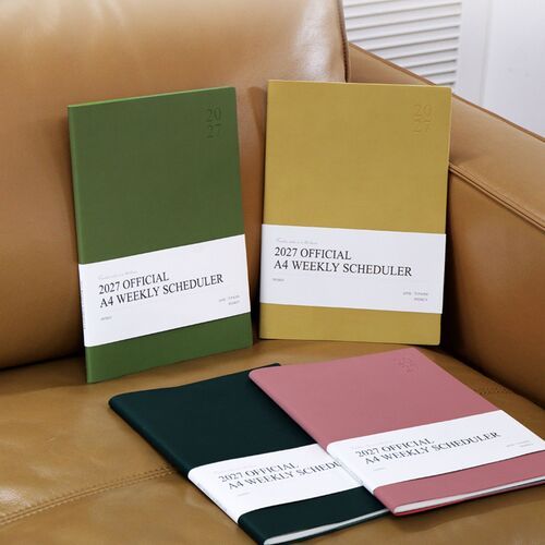 Green Mousse and Mustard schedulers stand upright against a warm chair, displaying their understated embossed 2027 covers.