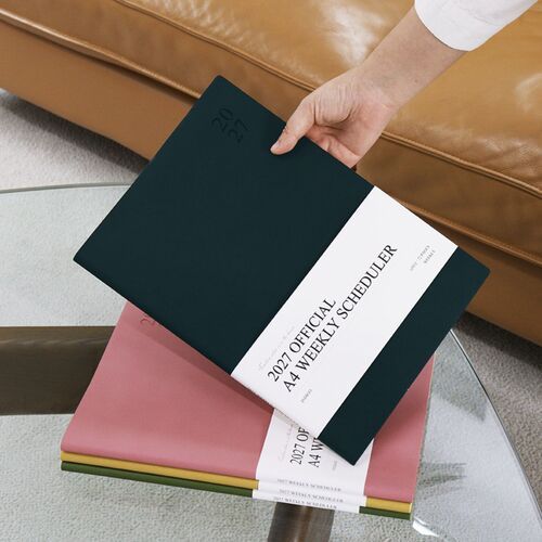A stack of Blossom, Deep Forest, and Green Mousse schedulers rests on a chair, showing their slim A4 profiles.