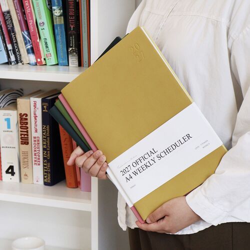 Green Mousse scheduler is held beside a laptop, showing how the large A4 format suits desk planning.
