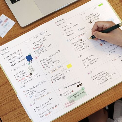 A Mustard scheduler is held in front of a bookshelf, highlighting its clean cover and wide portrait format.