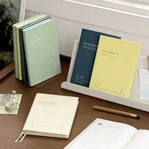 Four 2027 The Planner M styles are arranged on a desk, showing Milk White, Butter, Cream Mint, and Steel Blue covers.
