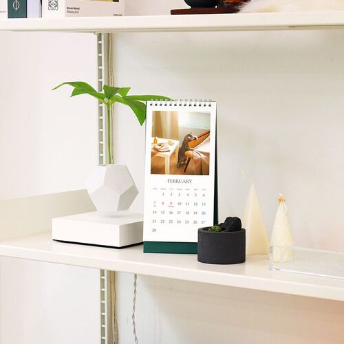 A February page is displayed on a shelf with decor, demonstrating the calendar’s upright triangular support in a narrow space.