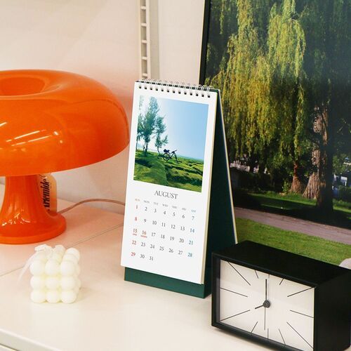 August is displayed beside an orange lamp and framed art, showing the calendar as a workspace accent with a scenic photo.