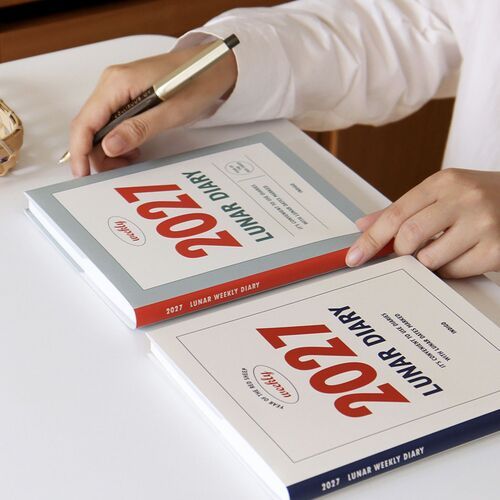 An open planner rests on a desk while a hand writes, showing its practical format for everyday notes and scheduling.