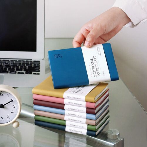 Teal Blue is lifted above a neat stack of colorful planners beside a laptop and desk clock.