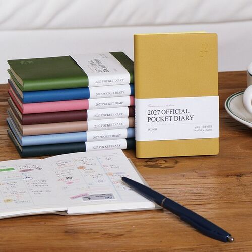 Mustard stands upright beside a stack of planners, with an open monthly page and pen arranged nearby.