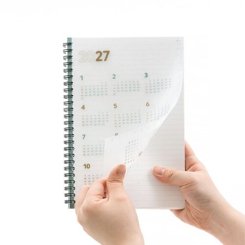 A hand lifts the translucent PP cover to reveal the yearly calendar and lined page beneath it.