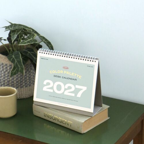 The 2027 cover is shown on a desk beside a plant and mug, emphasizing the pale blue palette and spiral-bound construction.