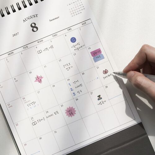 The scheduler sits on stacked books beside stationery, making a tidy addition to a reading or work corner.