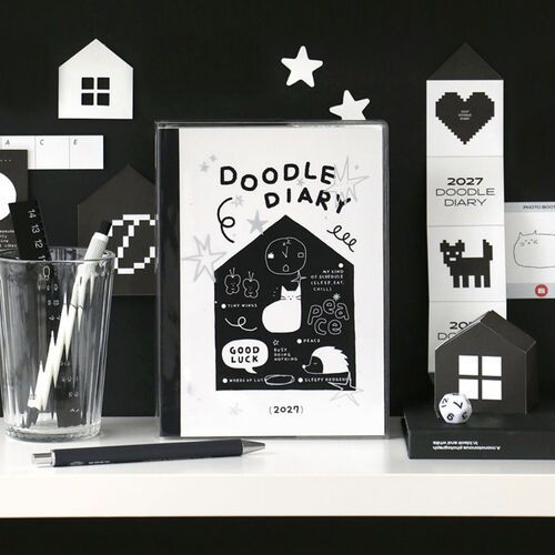 The White planner stands upright beside stationery, displaying its illustrated cover and black spine in a desk setting.