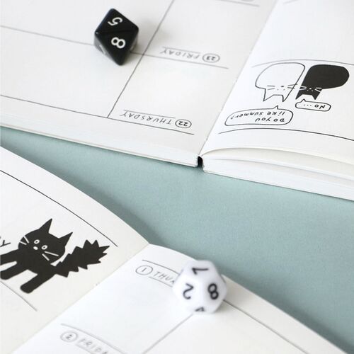 A White planner lies over an open schedule with its cat illustration, clear cover, and black spine visible.