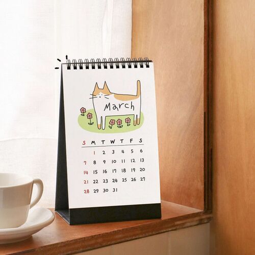 Photo 9: A March page sits on a black stand near a cup, showing the calendar’s upright format and seasonal cat illustration.
