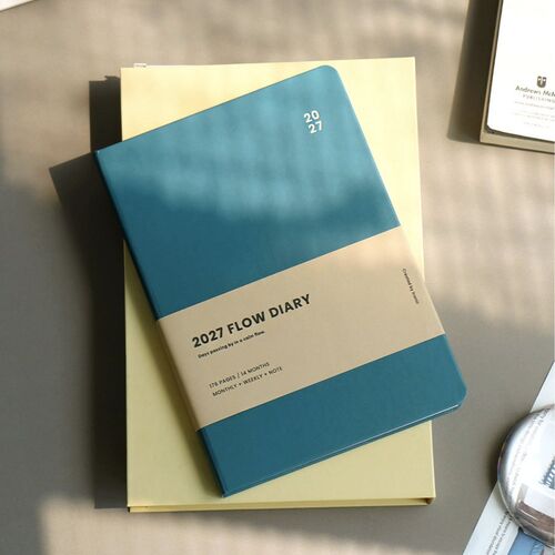 Teal Blue rests over pale paper in a sunlit setting, with the 2027 cover label clearly visible.