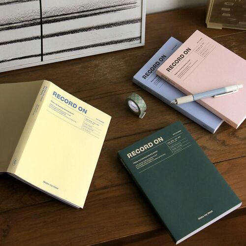 Several closed planners are arranged on a wooden table, showing the slim profile and four available cover colors.